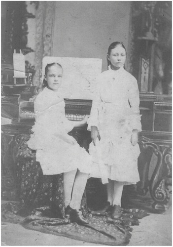 Studio portrait of Olive Fremstad standing in front of a piano at which is seated one of her pupils.