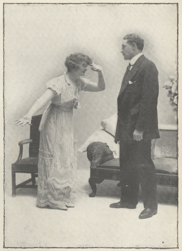 Miss Billie Burke as Lily stands in front of Mr. Herbert as Captain Jeyes with one arm outstretched and the index finger of the other arm pointing to her temple.