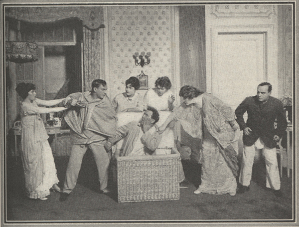 Photograph of several actors and actresses gather around an actor who is emerging from hiding in a wicker basket.