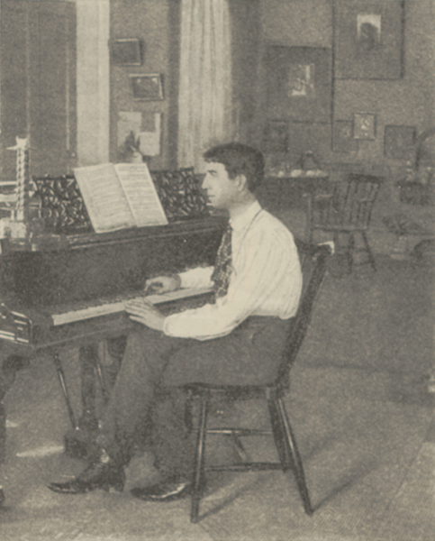 Ethelbert Nevin seated on a cane-back chair at a piano, looking at the music with his hands on the keys of the piano