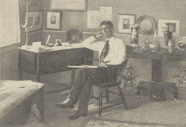 Ethelbert Nevin seated in his studio in a cane-back chair in front of a desk, resting his elbow on the desk and holding papers in his lap