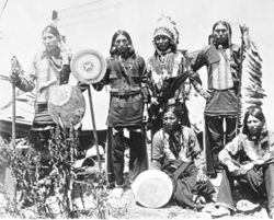 Six Native American men in traditional regalia in front of tipi