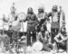 Six Native American men in traditional regalia in front of tipi