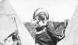 Native American man wearing headdress and blanket standing between tipis